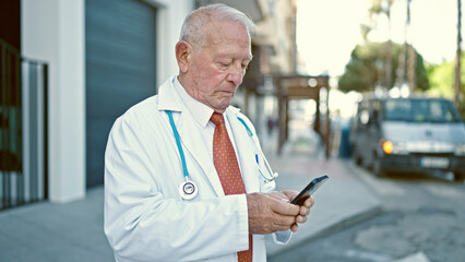 Senior grey-haired man doctor standing with serious expression using smartphone at street