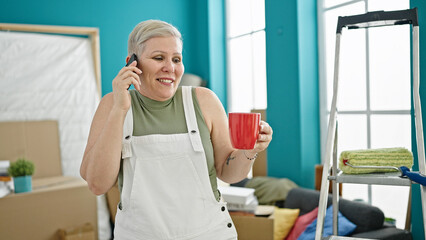 Middle age grey-haired woman talking on smartphone drinking coffee at new home