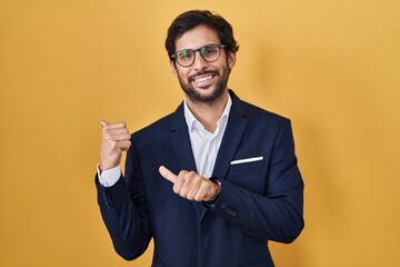 Handsome latin man standing over yellow background pointing to the back behind with hand and thumbs...