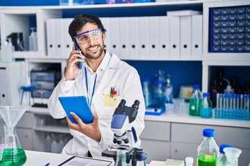 Young hispanic man scientist talking on the smartphone using touchpad at laboratory