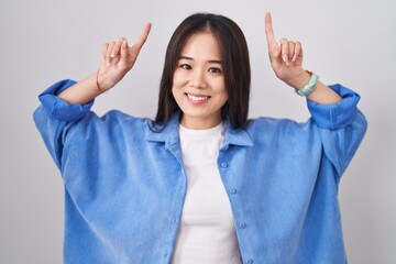 Young chinese woman standing over white background smiling amazed and surprised and pointing up with fingers and raised arms.