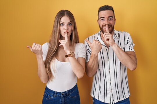 Young couple standing over yellow background asking to be quiet with finger on lips pointing with hand to the side. silence and secret concept.