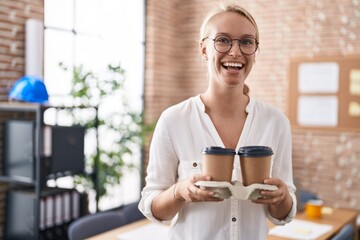 Young caucasian woman working at the office holding coffee cups smiling and laughing hard out loud because funny crazy joke.