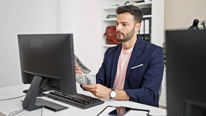 Fototapeta premium Young hispanic man business worker using computer counting dollars at office