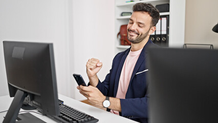 Young hispanic man business worker using computer and smartphone with winner gesture at office