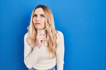 Young caucasian woman standing over blue background thinking concentrated about doubt with finger...