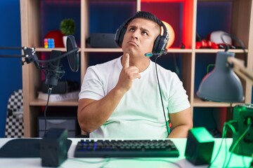 Young hispanic man playing video games thinking concentrated about doubt with finger on chin and looking up wondering