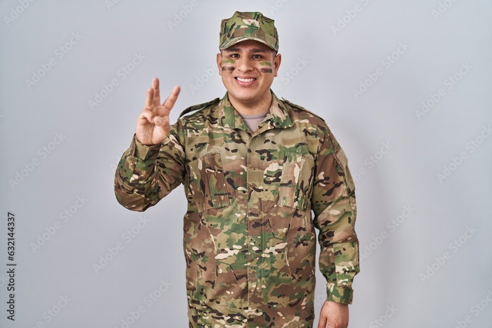 Canvas Prints Hispanic young man wearing camouflage army uniform showing and pointing up with fingers number three while smiling confident and happy.