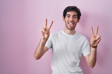 Young hispanic man standing over pink background smiling with tongue out showing fingers of both hands doing victory sign. number two.