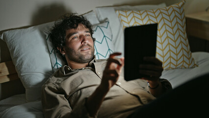 Young hispanic man using touchpad lying on bed at bedroom