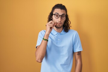 Young hispanic man standing over yellow background mouth and lips shut as zip with fingers. secret...