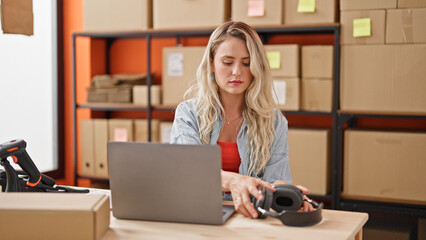 Young blonde woman ecommerce business worker using laptop holding headphones at office
