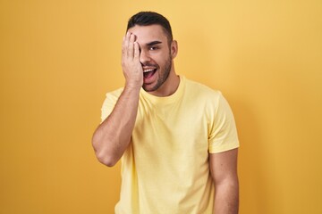 Young hispanic man standing over yellow background covering one eye with hand, confident smile on face and surprise emotion.