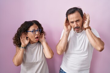 Middle age hispanic couple together over pink background trying to hear both hands on ear gesture, curious for gossip. hearing problem, deaf