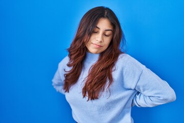 Obraz premium Hispanic young woman standing over blue background suffering of backache, touching back with hand, muscular pain