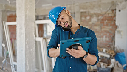 Young hispanic man architect using touchpad device speaking on the phone at construction site