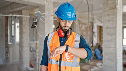 Young hispanic man builder looking at wrist watch at construction site