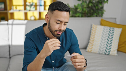 Young hispanic man thinking about money holding credit card at home