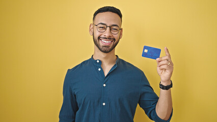 Young hispanic man smiling confident holding credit card over isolated yellow background