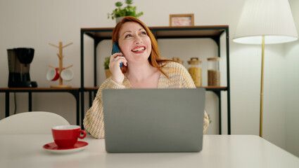 Young redhead woman using laptop and talking on the smartphone at home