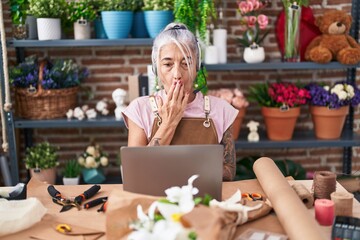 Middle age woman with tattoos working at florist shop doing video call covering mouth with hand, shocked and afraid for mistake. surprised expression