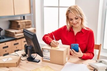 Young blonde woman ecommerce business worker writing on package using smartphone at office