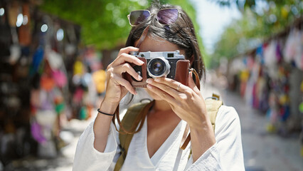 Young beautiful hispanic woman tourist using vintage camera at street market