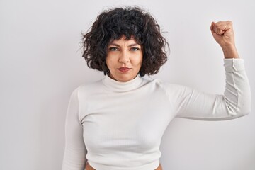 Hispanic woman with curly hair standing over isolated background strong person showing arm muscle, confident and proud of power