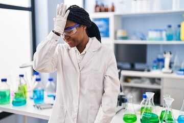 African american woman working at scientist laboratory surprised with hand on head for mistake, remember error. forgot, bad memory concept.