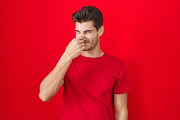 Young hispanic man standing over red background smelling something stinky and disgusting, intolerable smell, holding breath with fingers on nose. bad smell