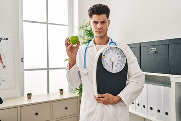 Young hispanic doctor man holding scale at dietitian clinic skeptic and nervous, frowning upset...