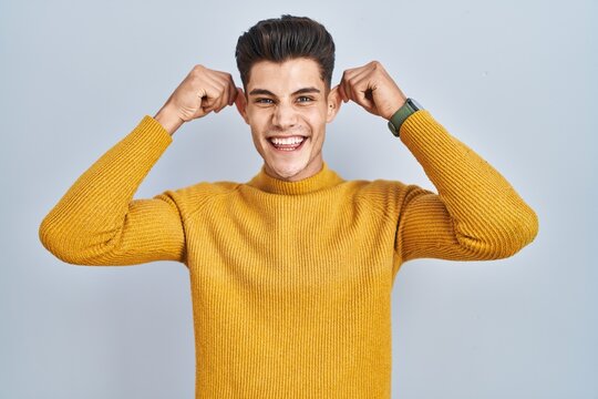 Young hispanic man standing over blue background smiling pulling ears with fingers, funny gesture. audition problem