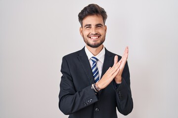 Young hispanic man with tattoos wearing business suit and tie clapping and applauding happy and joyful, smiling proud hands together