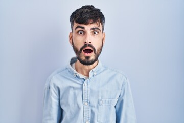 Young hispanic man with beard standing over blue background afraid and shocked with surprise and amazed expression, fear and excited face.