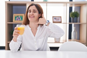 Fototapeta premium Brunette woman drinking glass of orange juice smiling doing phone gesture with hand and fingers like talking on the telephone. communicating concepts.