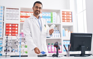 Young hispanic man pharmacist holding pills using credit card and dataphone at pharmacy