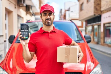 Young hispanic man with beard wearing delivery uniform and cap holding dataphone clueless and...