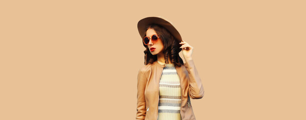 Portrait of beautiful young woman wearing jacket, sunglasses, round hat on brown wall studio background