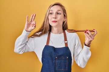 Young blonde woman wearing apron tasting food holding wooden spoon smiling looking to the side and staring away thinking.