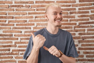 Young caucasian man standing over bricks wall pointing to the back behind with hand and thumbs up, smiling confident