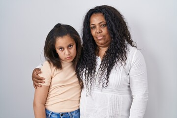 Mother and young daughter standing over white background relaxed with serious expression on face. simple and natural looking at the camera.