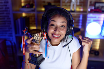 Young arab woman playing video games holding trophy screaming proud, celebrating victory and success very excited with raised arm