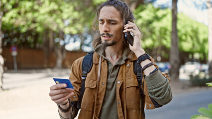Young hispanic man tourist talking on smartphone holding credit card at park