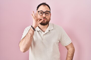 Plus size hispanic man with beard standing over pink background mouth and lips shut as zip with...
