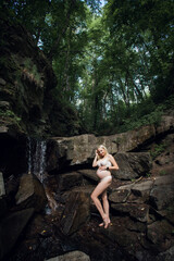 A smiling happy pregnant girl in light underwear, with blond short hair, stands against the backdrop of picturesque rocks near a waterfall in the forest.
