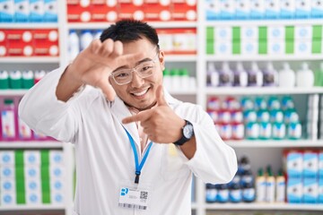 Chinese young man working at pharmacy drugstore smiling making frame with hands and fingers with happy face. creativity and photography concept. © Krakenimages.com