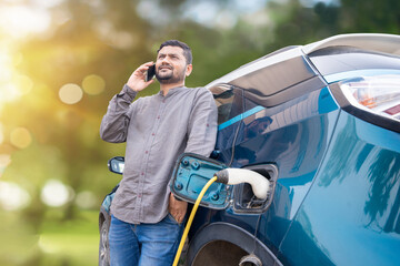 Happy Indian man talking on phone while his modern electric car getting charge at charging station....