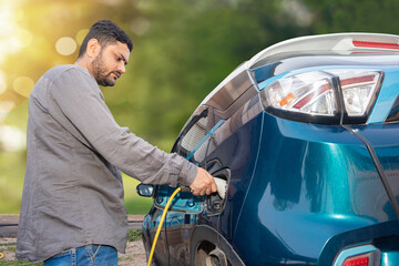 Indian man attaching power cable into socket to charge his modern electric car at outdoor charging station. Electric vehicle Recharging battery charging port. Renewable energy or green energy.