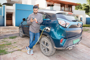 Plakat Happy Indian man using smartphone while his modern electric car getting charge outside house. Electric vehicle Recharging battery charging port. Renewable energy or green energy. Full length.