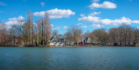 A lakeside restaurant pavilion in the Hirschgarten park, Munich, Germany, surrounded by leafless trees under a blue sky with clouds. A scenic city park landscape for travel and leisure themes.